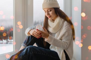 young-attractive-woman-stylish-white-knitted-sweater-scarf-hat-sitting-home-windowsill-christmas-holding-glass-snow-ball-present-decoration-winter-forest-view-lights-bokeh
